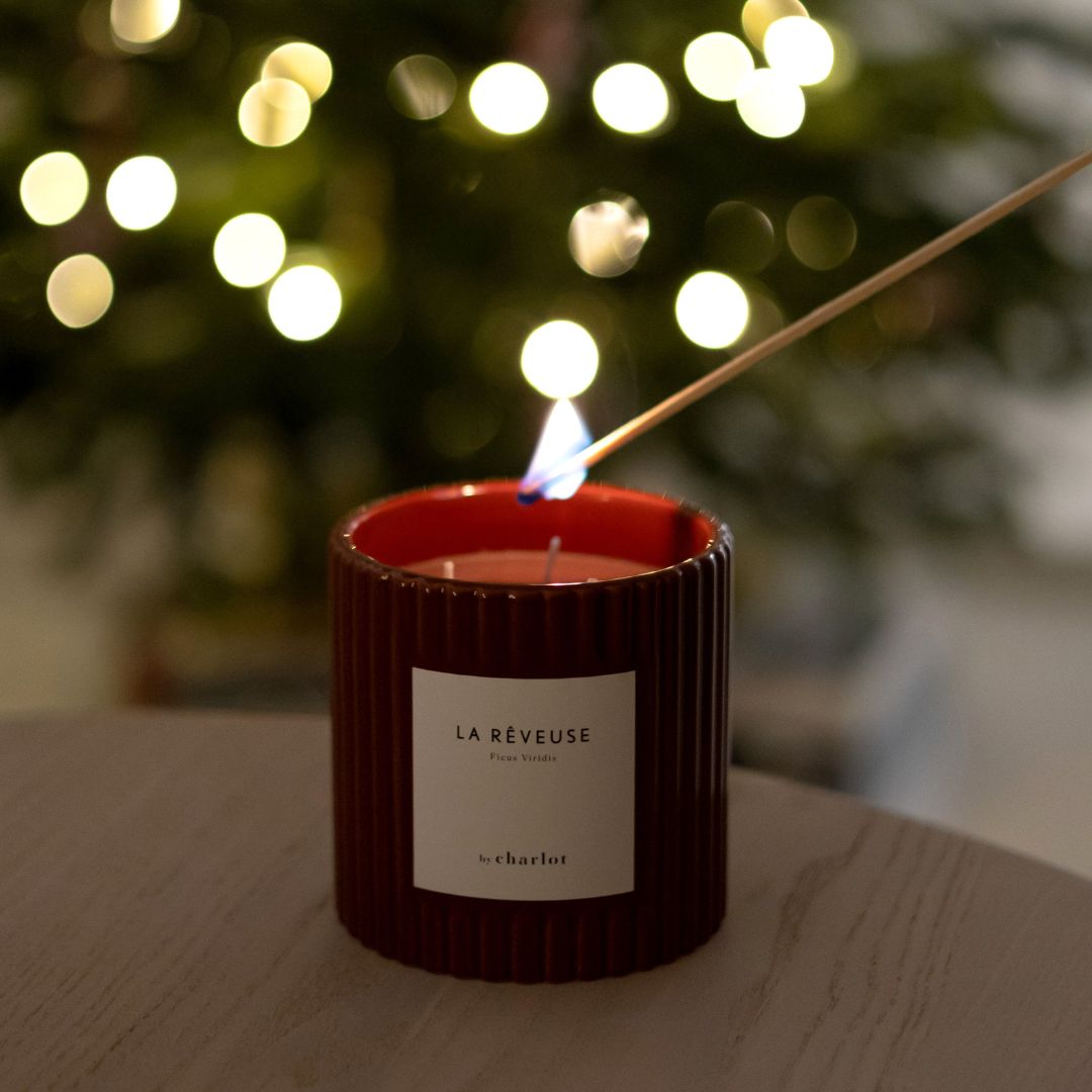 Bougie parfumée La Rêveuse By Charlot allumée avec une longue allumette, pot en céramique cannelée rouge, sapin illuminé en arrière-plan
